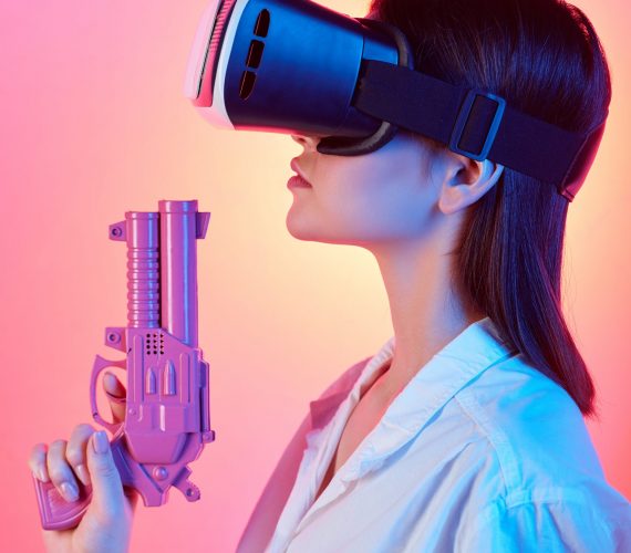 Pretty girl in vr headset holding violet plastic gun while shooting at virtual target against pink neon background in isolation